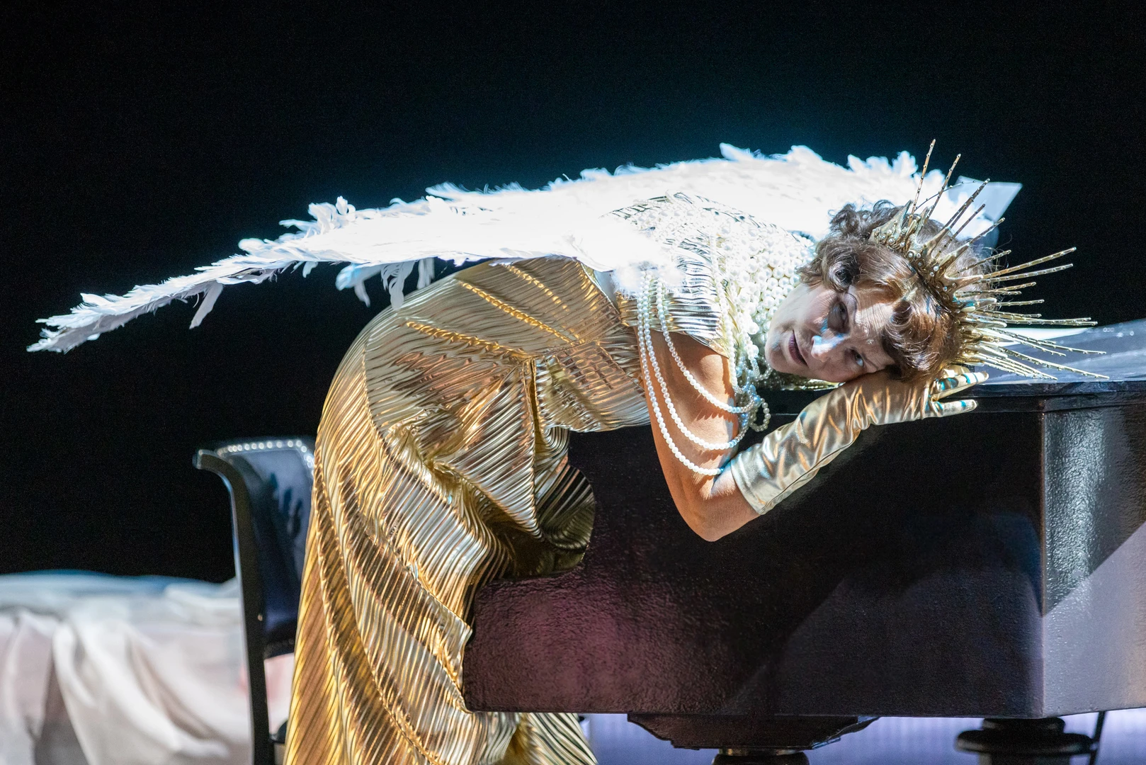 A person in a gold-coloured costume with large white wings is leaning over a wing on a dark stage. Bright light makes the figure stand out against the dark background.