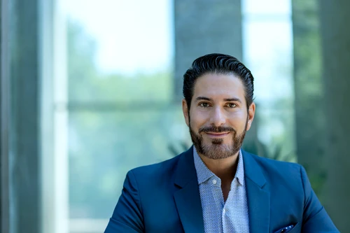 Mann mit dunklen Haaren und dunklem Dreitagebart in dunkelblaumen Anzug mit hellem Hemd vor einem Fenster