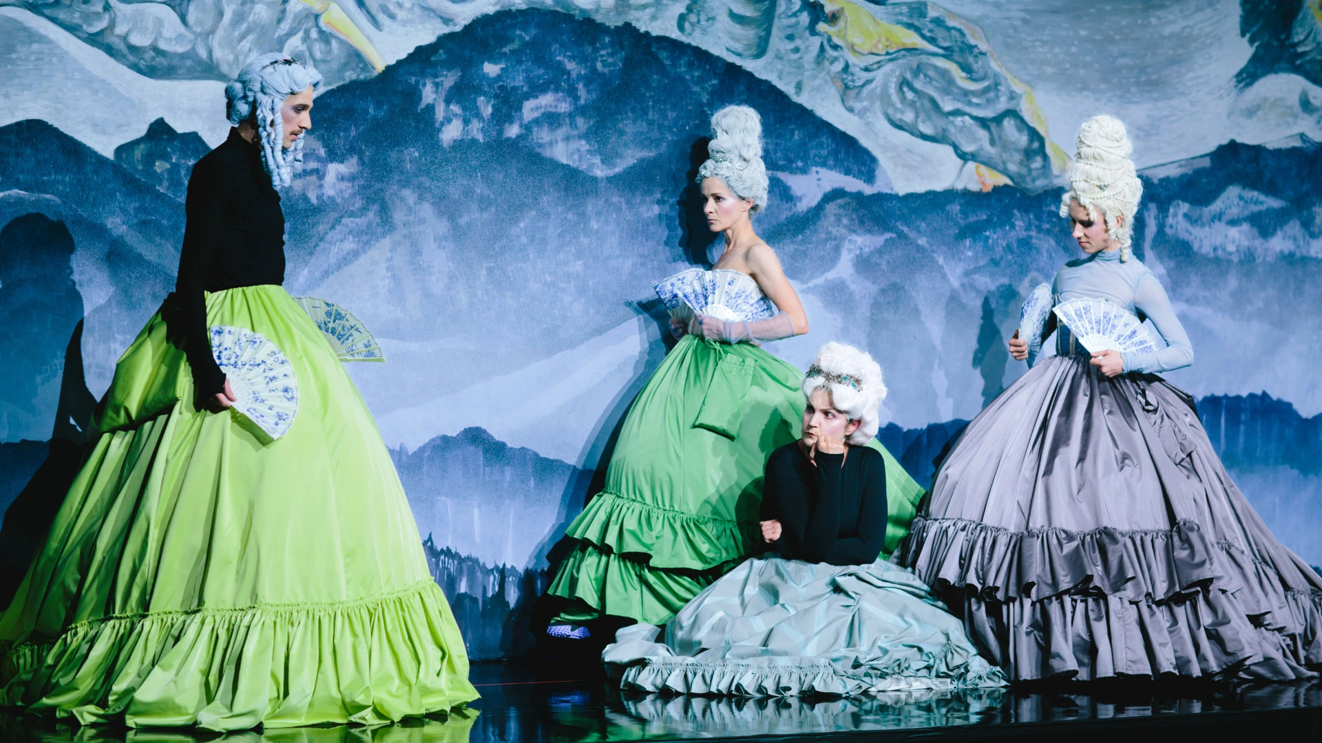 Vier Darstellerinnen in barock inspirierten Kostümen mit Perücken und Fächern auf einer Theaterbühne.