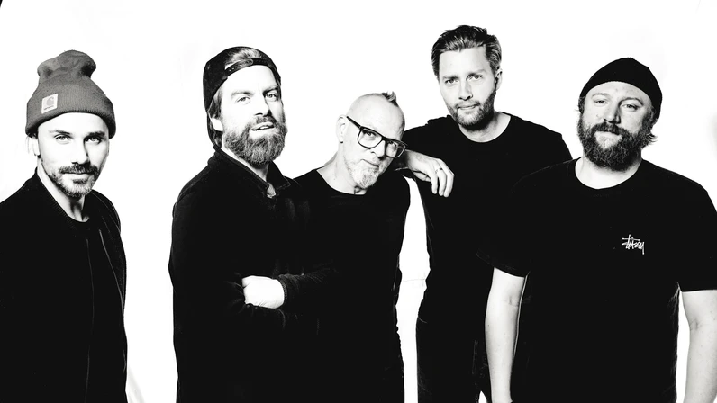 Thomas D und die KBCS stehen in dunkler Kleidung vor weißem Hintergrund eng beisammen, wobei zwei eine Mütze tragen, einer eine umgedrehte Kappe trägt und einer Brille und grauen Bart hat, und alle mit ruhigen, freundlichen Blicken in die Kamera schauen.