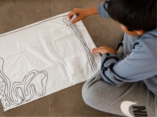 A child is sitting on the floor, we can only see the side he is facing away from. He is drawing with a black pen on a white cushion cover. 