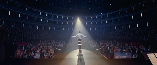 Eine Person von Hinten auf einer Bühne, dahinter ein großer Saal schwach beleuchtet voller Publikum