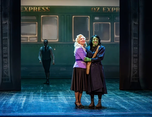 Blonde Frau mit schwarzem Rock und lavendelfarbigem Sakko und schwarzhaarige Frau mit grüner Haut in schwarzem Mantel stehen lächelnd in Umarmung, im Hintergrund ein Zug mit dem Schriftzug "Oz Express"