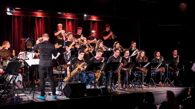 Eine große Jazz‑Bigband mit jungen Musikerinnen und Musikern spielt auf einer Bühne unter warmem Scheinwerferlicht, während Andreas Pranzl als Dirigent vorne steht und die Gruppe anleitet.
