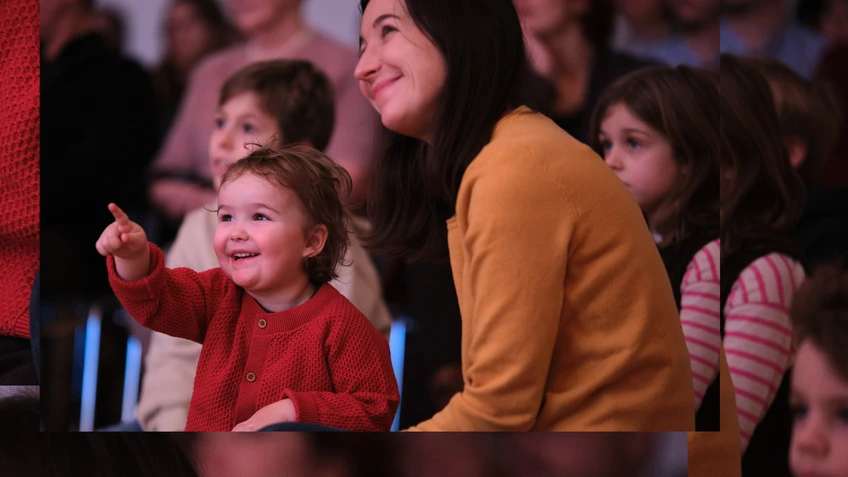 Audience photo from the ‘Tonspiele’ project ‘Schnee’ (Snow) – a project of the music education programme of the Tonkünstler Orchestra Lower Austria. The photo shows happy children listening and watching intently. A child sits on his mother's lap and points towards the stage.