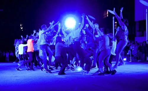 Ein Bühnenbild von einer Gruppe an Menschen in blauen Scheinwerferlicht, die Hände sind nach oben gestreckt.