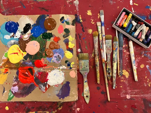 Farbpalette mit bunten Acrylfarben, daneben mehrere farbbespritzte Pinsel und eine Schachtel mit Pastellkreiden auf einer kreativ genutzten Arbeitsfläche.