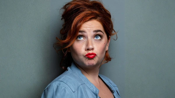 Rothaarige Frau mit rotem Lippenstift schürzt die Lippen und dreht die Augen nach oben Richtung Haare