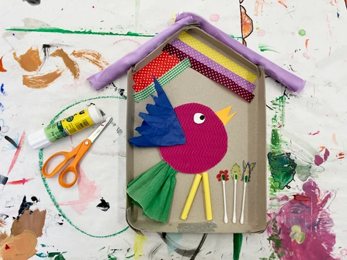 Bunte Bastelarbeit in Form eines vogelförmigen Motivs, gestaltet aus Papier, Stoffresten und kleinen Materialien in einem kartonähnlichen Rahmen. Daneben liegen eine Schere und ein Klebestift auf einer farbverschmierten Unterlage.