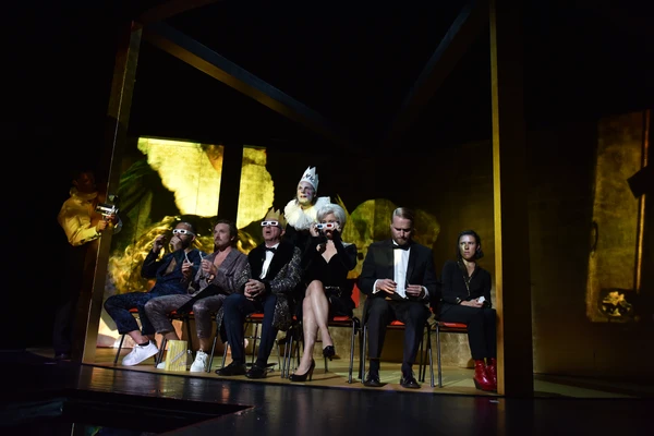 Szenenfoto aus "Hamlet". Sechs Schauspieler*innen sitzen mit dem Rücken zum König, ein paar von ihnen tragen 3D-Brillen.