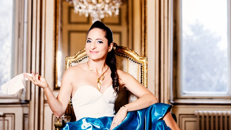 Caroline Athanasiadis, elegant gekleidet, sitzt in einem prunkvollen Saal auf einem goldenen Stuhl, trägt ein weißes Kleid mit blauem Rock und roten High Heels, hält ein Taschentuch in der Hand und lächelt selbstbewusst vor einem großen Kronleuchter.