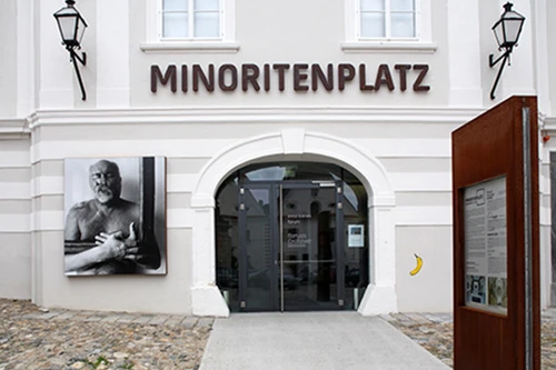 Eingang am Minoritenplatz mit Bogenportal; links ein großformatiges Schwarz-Weiß-Porträt, darüber der Schriftzug MINORITENPLATZ.