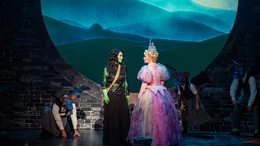 Green-skinned woman with black hair and black clothing and blonde woman in an exaggerated pink princess dress and huge tiara, facing each other as if in conversation, masked figures in the shadows, projections of a misty hilly landscape in the background