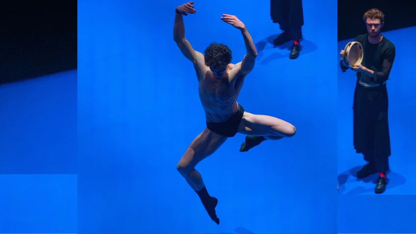 Tänzer:innen, schwarz gekleidet, auf einer auffällig blauen Bühne, einer von ihnen lediglich in schwarzer Unterhose, gezeigt in einer Sprungfigur, neben ihm ein Performer in schwarzem langärmligen Shirt, schwarzem Rock, ein Tambourin in der Hand.