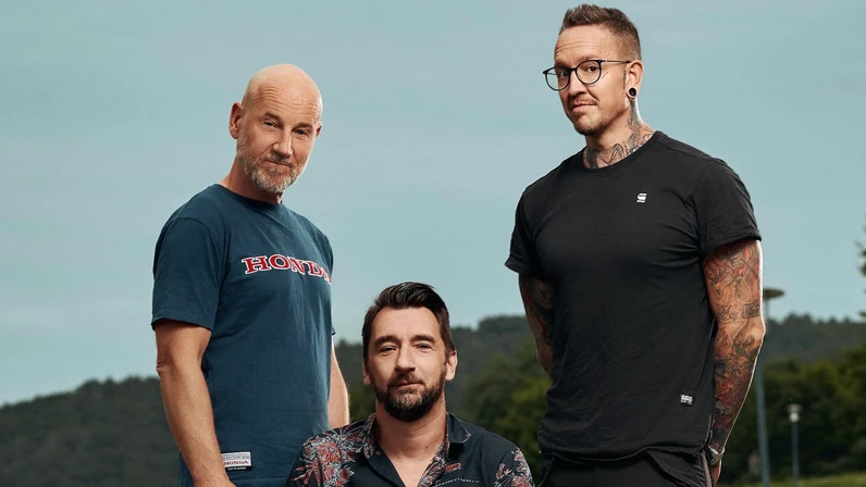 Roland Düringer, Bernhard Speer und Bernhard Egger posieren auf einer Landstraße vor bewaldeten Hügeln, wobei Egger in einem roten Sessel sitzt und die beiden anderen locker daneben stehen.