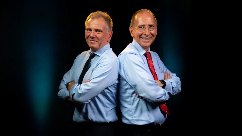 Armin Wolf und Peter Filzmaier stehen in hellblauen Hemden Rücken an Rücken vor dunklem Hintergrund, lächelnd mit verschränkten Armen, wobei einer eine rote Krawatte und der andere eine dunkle Krawatte trägt.