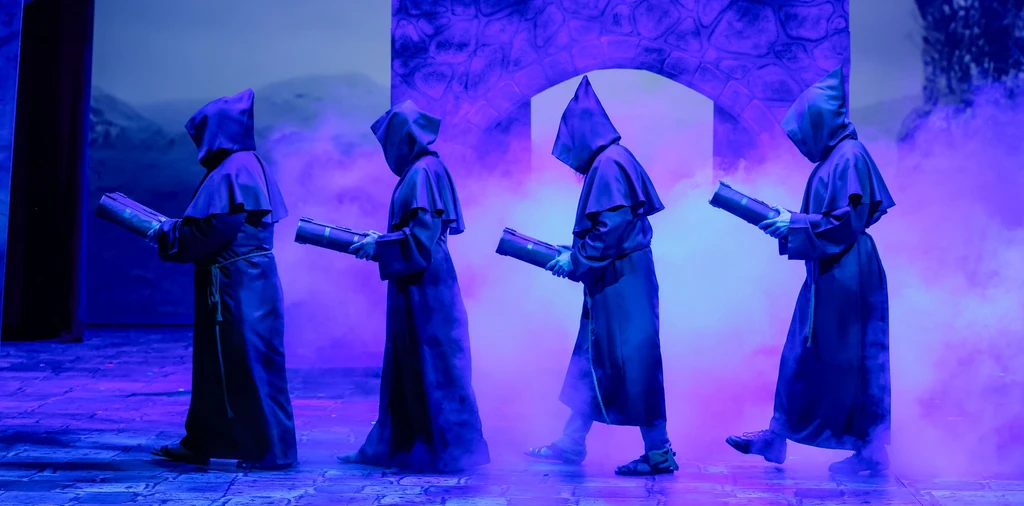 Vier Mönche mit großen Büchern in der Hand sind von der Seite in blauem Bühnenlicht zu sehen. Im Hintergrund ist Nebel und ein Mauertor.