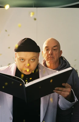 Mann mit schwarzer Haube bläst Glitzer aus einem großen aufgeschlagenen Buch, ein anderer Mann sieht ihm über die Schulter