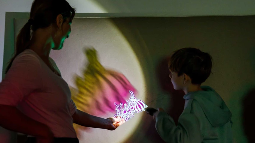 Artist Iva Ivanova holds a plexiglass silhouette through which a boy shines a torch, casting a colourful shadow.