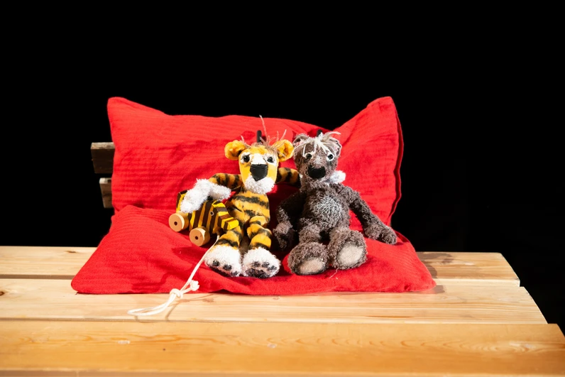Zwei weiche Tierpuppen – ein Tiger mit Tigerente an der Hand und ein Bär – sitzen eng nebeneinander auf einem roten Kissen auf einem Holztisch vor schwarzem Hintergrund.