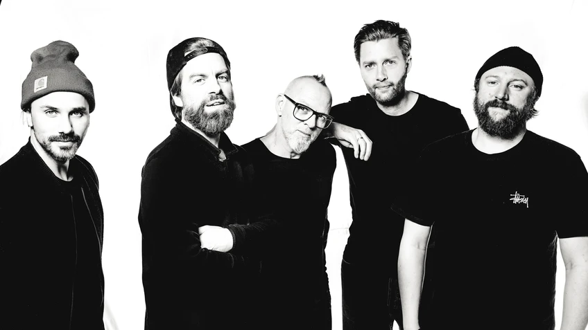 Thomas D und die KBCS stehen in dunkler Kleidung vor weißem Hintergrund eng beisammen, wobei zwei eine Mütze tragen, einer eine umgedrehte Kappe trägt und einer Brille und grauen Bart hat, und alle mit ruhigen, freundlichen Blicken in die Kamera schauen.