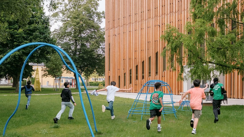 Children run across a green meadow towards the wooden building of the KinderKunstLabor.