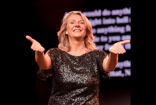 Eine Frau in schwarz glitzerndem Kleid lächelt ins Publikum und streckt die Arme einladend aus