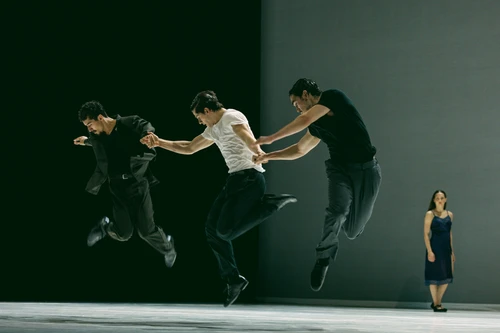 Szenenfoto von "Obsidian", einer Choreografie von Bobbi Jene Smith und Or Schraiber. Zu sehen sind Tänzer:innen des Ballet BC, drei von ihnen im Bildvordergrund, die gleichzeitig hochspringen, im Hintergrund steht eine Tänzerin im schwarzen Kleid vor dem grauen Bühnenhintergrund.