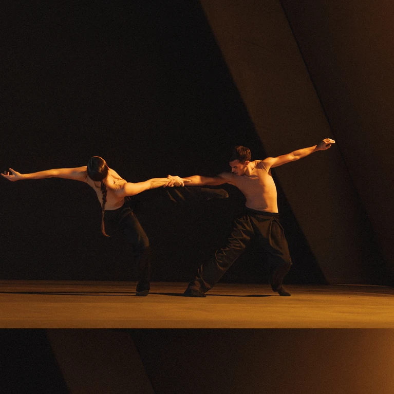 A performance photo of the choreography ‘SWAY’ by Medhi Walerski. It shows two dancers from Ballet BC, dressed in black trousers and white shirts, against a black stage backdrop, performing a sweeping dance move, illuminated by yellow stage lighting. 