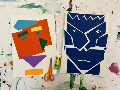 Zwei abstrakte Papier‑Porträts auf bemaltem Tisch, daneben Schere und Klebestift.