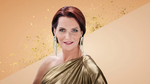 Porträtaufnahme von Maya Hakvoort in einem eleganten, einseitig drapierten, goldfarbenen Kleid mit auffälligen, langen Ohrringen. Der Hintergrund ist in warmen Beige- und Goldtönen gestaltet, mit dekorativen Glitzerelementen.