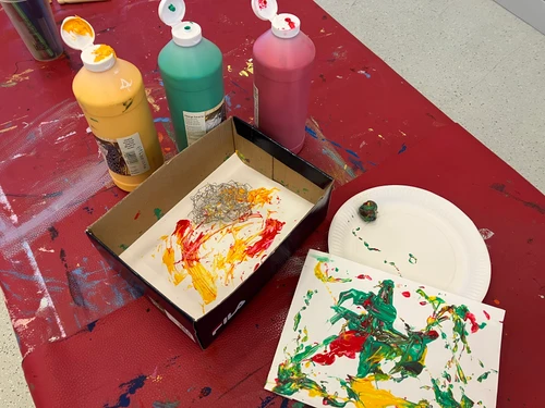 Basteltisch mit offenen Farbtuben in Gelb, Grün und Rot, einer Kartonschachtel mit bemaltem Papier und einer weiteren abstrakten Farbarbeit auf einem Teller.