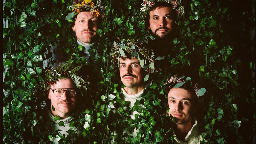 Fünf Männerköpfe mit Blumenkränzen ragen aus einer grünen Hecke