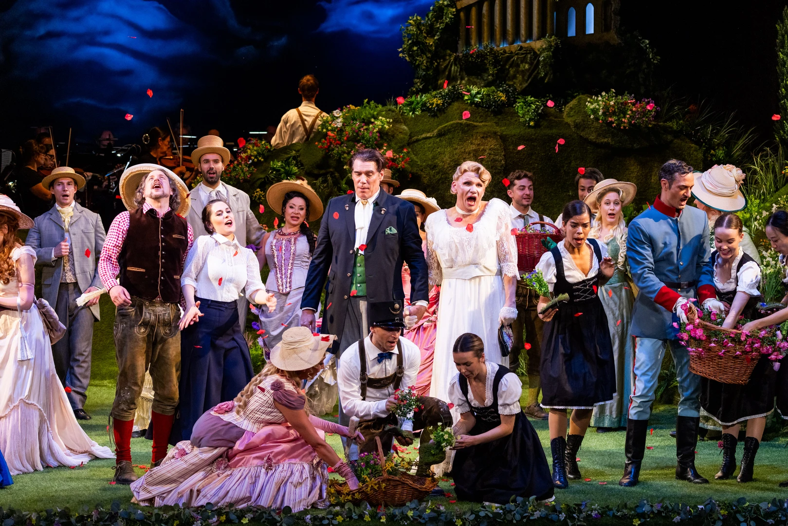 A large ensemble scene on a stage featuring numerous people in traditional dress. The group is arranged closely together and occupies a large part of the stage. Some figures are standing, whilst others are sitting or leaning slightly forwards. The costumes vary in colour and style, including long skirts, waistcoats, coats and hats. The background depicts a detailed, green landscape and a dark sky. The lighting clearly highlights the group at the front and ensures that all the figures are clearly visible.