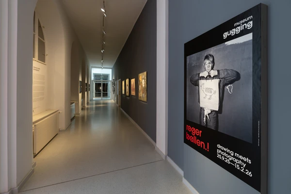 Moderner Ausstellungsflur mit beleuchteten Wänden, an denen großformatige Fotografien hängen; im Vordergrund ein Bild mit dunklem Hintergrund und weißer Schrift.