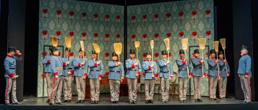 einige Männer in blauer Uniform mit roten Details nebeneinander gereiht mit Besen in der Hand, diese sind verkehrt nach oben gerichtet, Im Hintergrund Tapete mit Muster in türkis, rot und weiß